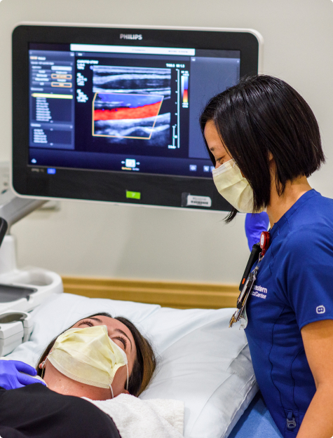 An ultrasonographer standing next to a patient’s bedside with the ultrasound monitor behind them