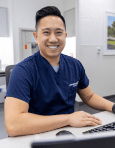Male healthcare professional in blue scrubs sitting within a hospital setting and smiling at the camera.