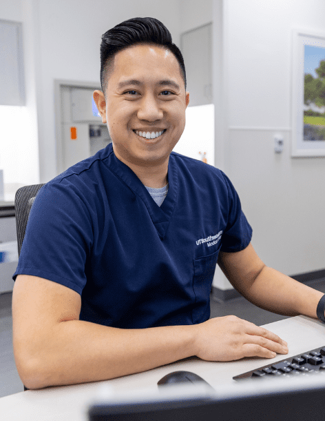 Male healthcare professional in blue scrubs sitting within a hospital setting and smiling at the camera.