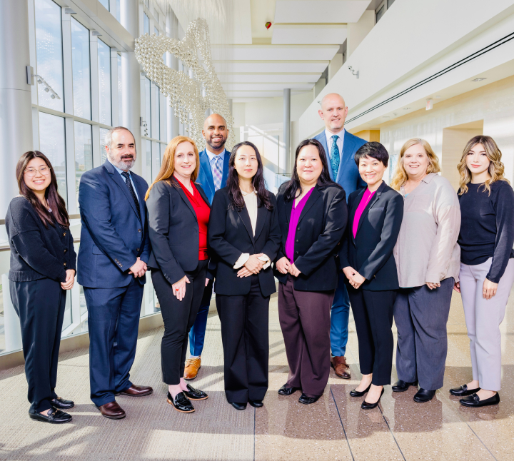 A group of nine professionals in business attire standing together in a brightly lit, modern indoor space with large windows in the background.
