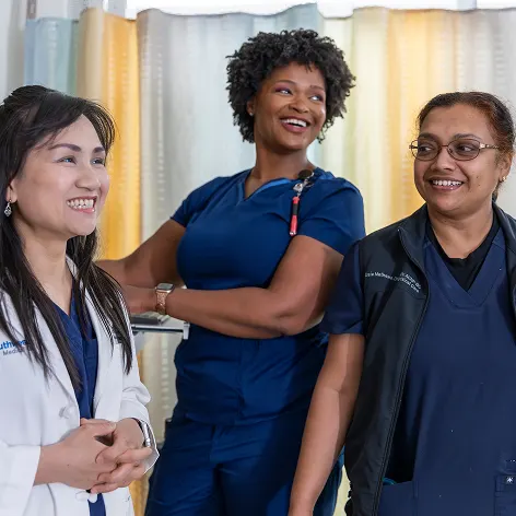Three female healthcare professionals in scrubs and a lab coat smile and converse in a clinical setting with a curtain in the background.